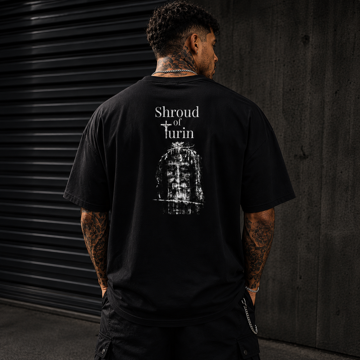 Oversized black Christian streetwear t-shirt featuring distressed Shroud of Turin back graphic worn by tattooed male model in luxury urban environment