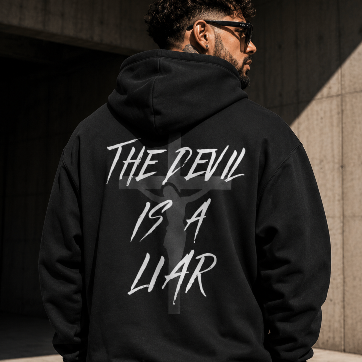 Back view of black Christian streetwear hoodie featuring “The Devil Is a Liar” graphic with faded cross design worn by male model outdoors.