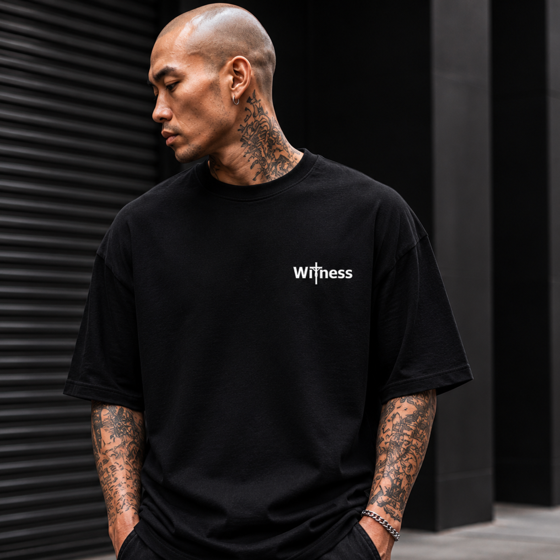 Premium oversized black Christian streetwear tee with minimalist Witness logo on front chest worn by tattooed male model